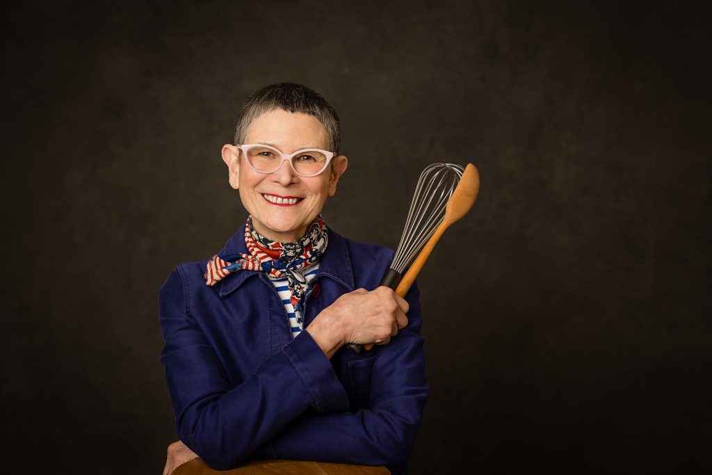 Photo of chef-author Dorie Greenspan holding a whisk and spoon against a black background